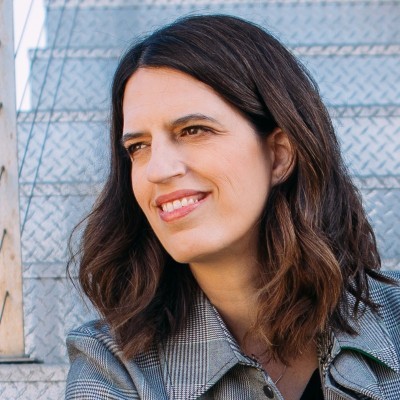 A woman with short curly hair wearing a black t-shirt looks into the camera.