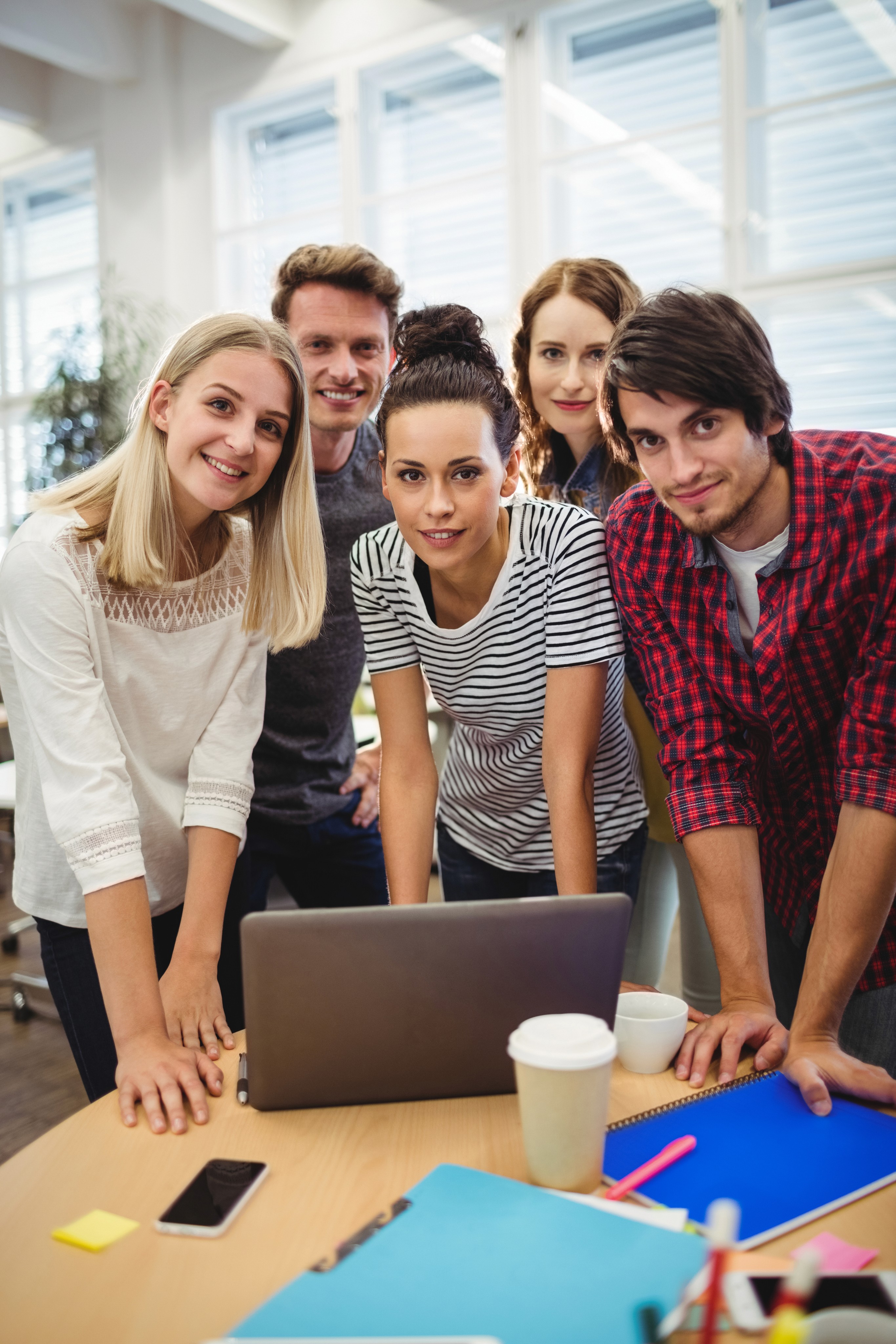 Five young adults gathered around a laptop in a bright office, smiling and leaning on a table with coffee cups and notebooks.