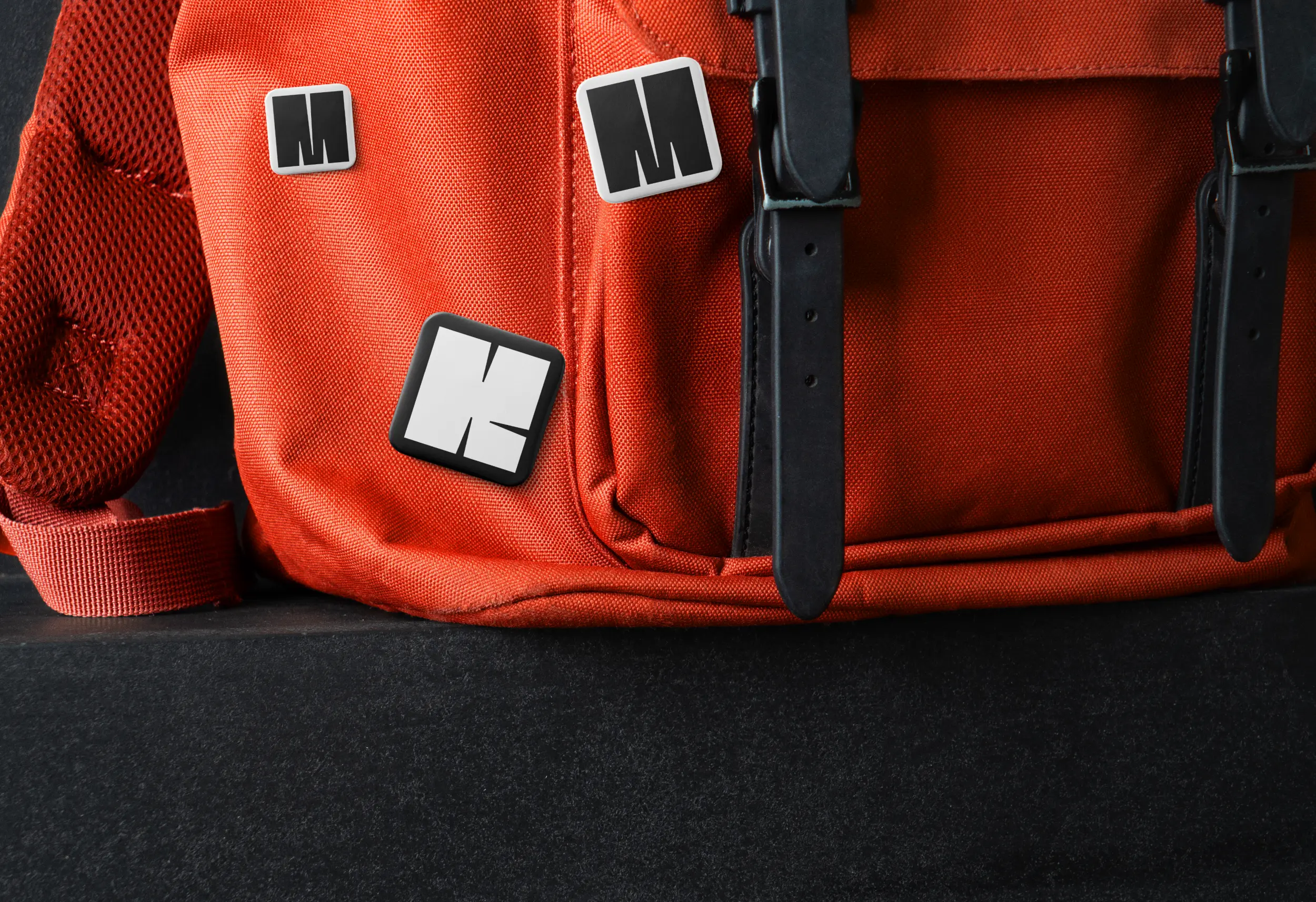 Orangefarbener Rucksack mit MMK-Ansteckern in Schwarz und Weiß auf dunkler Fläche