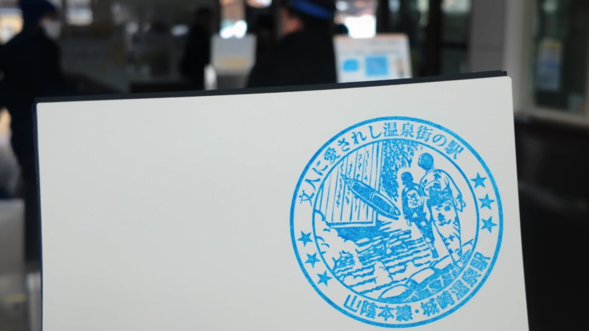 The Kinosaki Onsen station stamp printed in blue, showing bathers and a line of Japanese text.