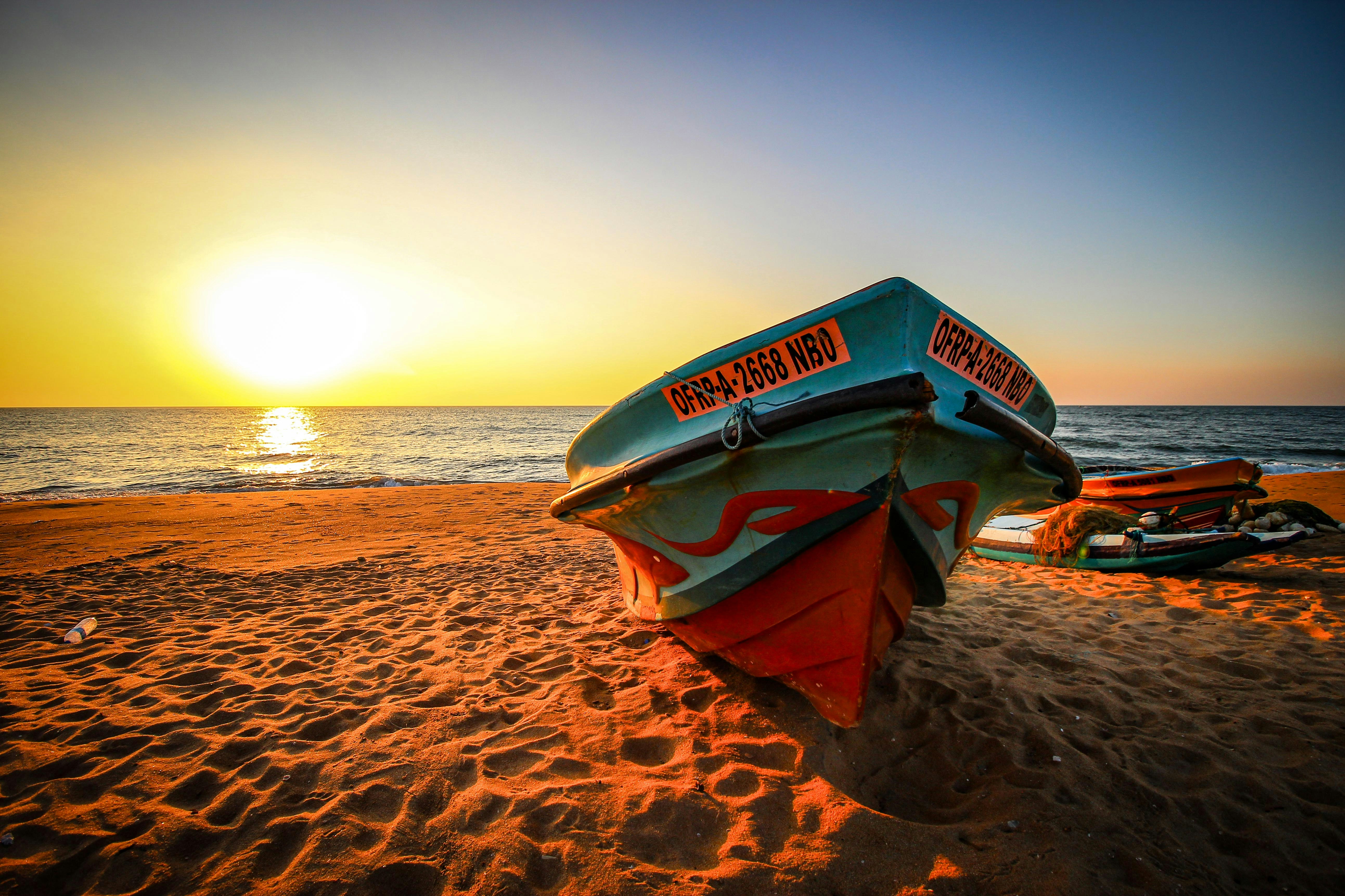 red and blue boat on brown sand near body of water during sunset