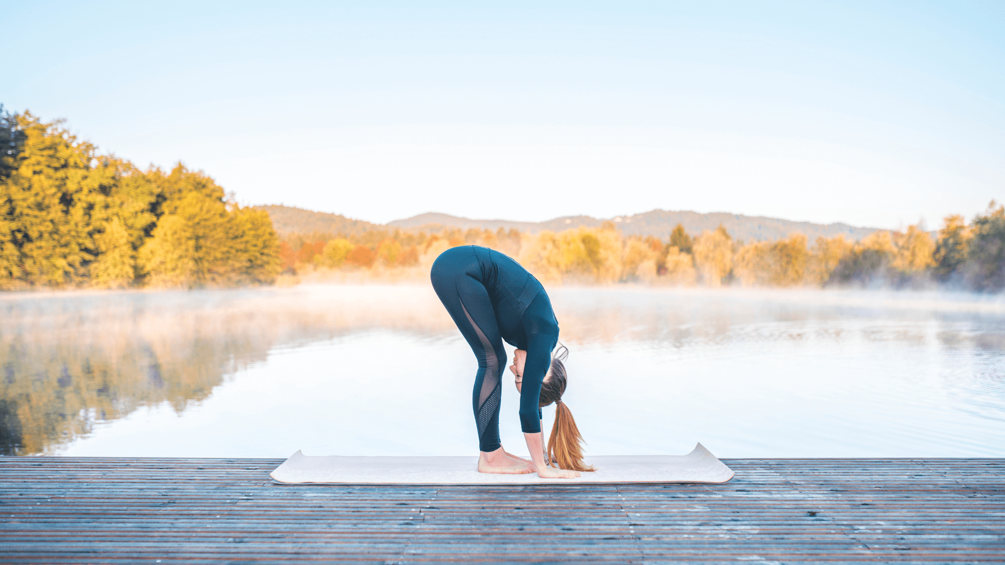 Woman practicing Standing Forward Fold (Uttanasana) on white yoga mat, on lakeside dock during misty autumn morning with fall foliage reflecting in calm water