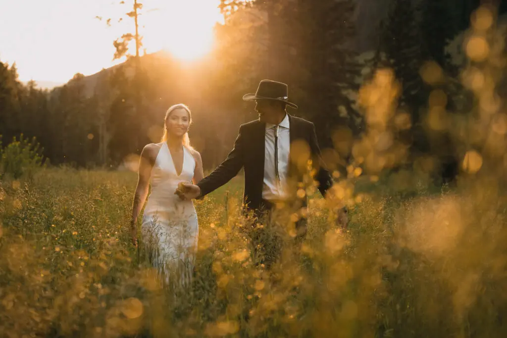 Romantic sunset wedding portrait of bride in halter dress and groom in cowboy hat walking through golden wildflower meadow with warm bokeh and mountain backdrop
