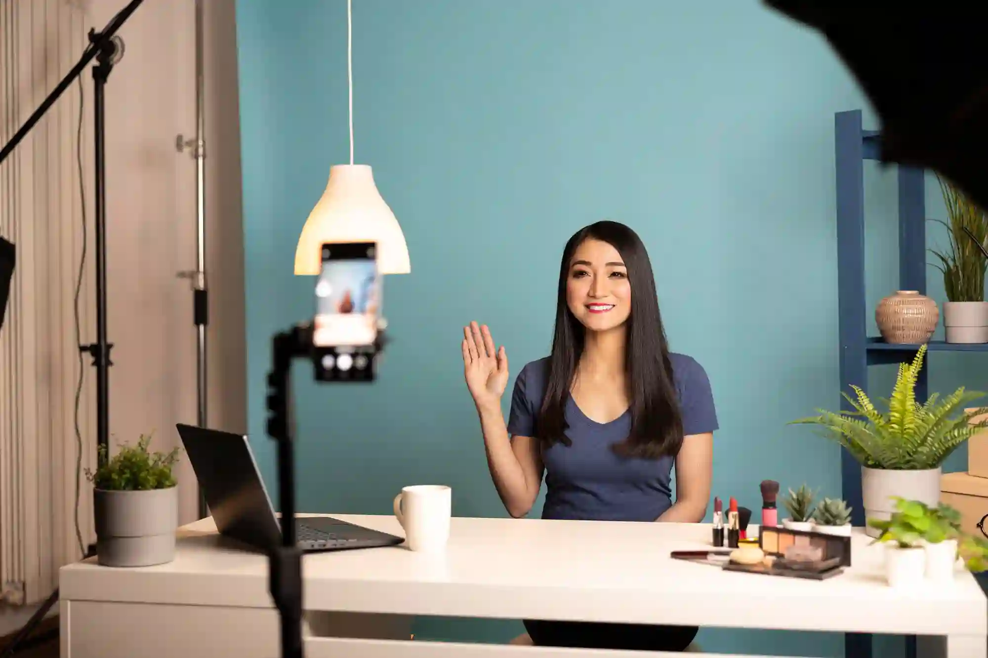 A woman waves at a smartphone camera set up for livestreaming.