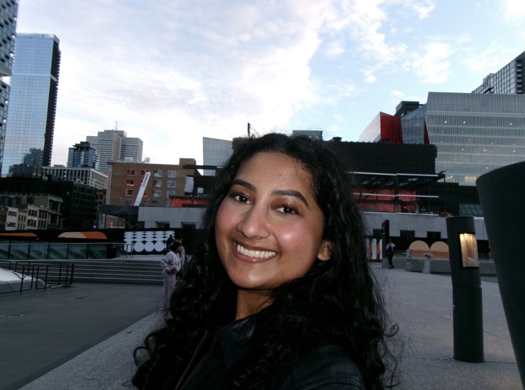 A person wearing headphones sits at a desk with a large curved monitor displaying graphic design software, surrounded by a laptop, smartphone, and potted plant, creating a modern workspace setup.