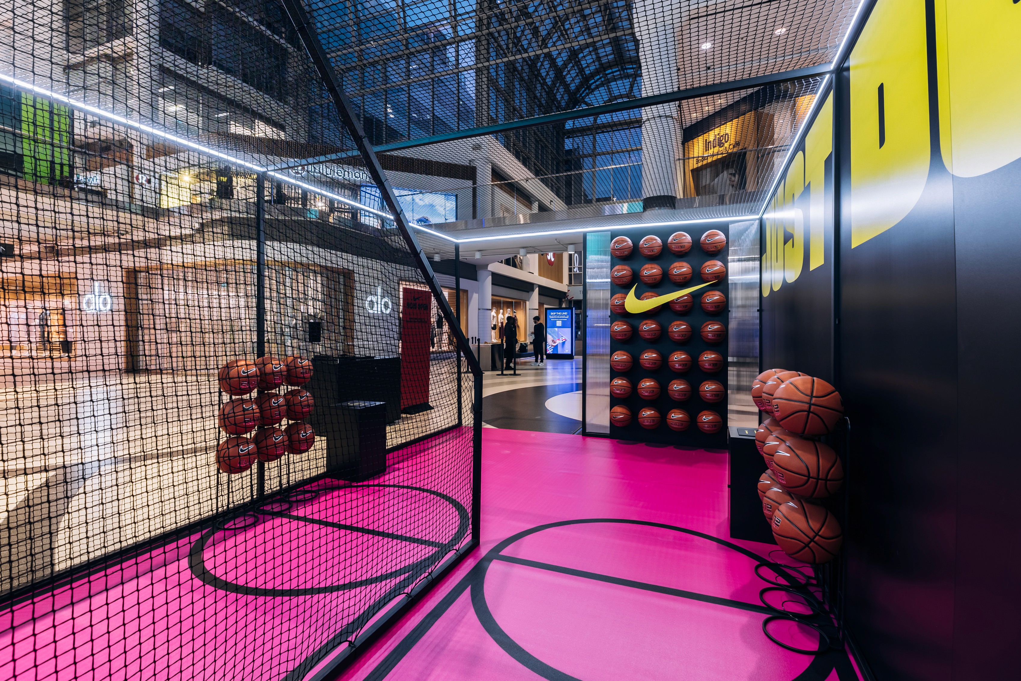 Indoor Nike-themed basketball-themed area in the Eaton Centre with pink flooring, basketballs on racks, and a black net enclosure. Bright, vibrant, energetic atmosphere.