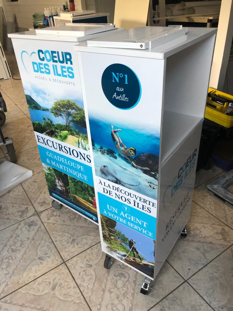 Deux stands promotionnels sur sol carrelé font la promotion d'excursions en Guadeloupe et en Martinique. Les images colorées représentent des paysages tropicaux et des explorations sous-marines. Les mots-clés incluent « Coeur des Iles » et « N°1 aux Antilles ».