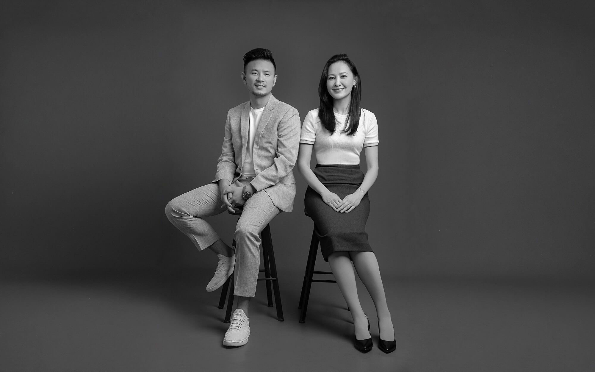 一男一女坐在高腳凳上,男士穿著淺色西裝和運動鞋,女士穿著白色上衣和深色半身裙,背景為純色牆面,黑白照片。
