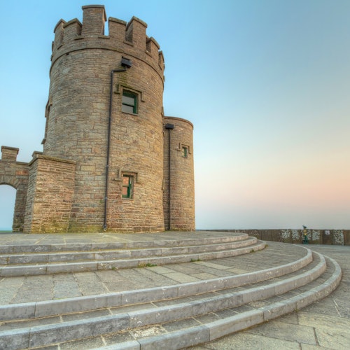 Кръгла каменна кула с бойници, прозорци и вход, заобиколена от извити стъпала под ясно небе.