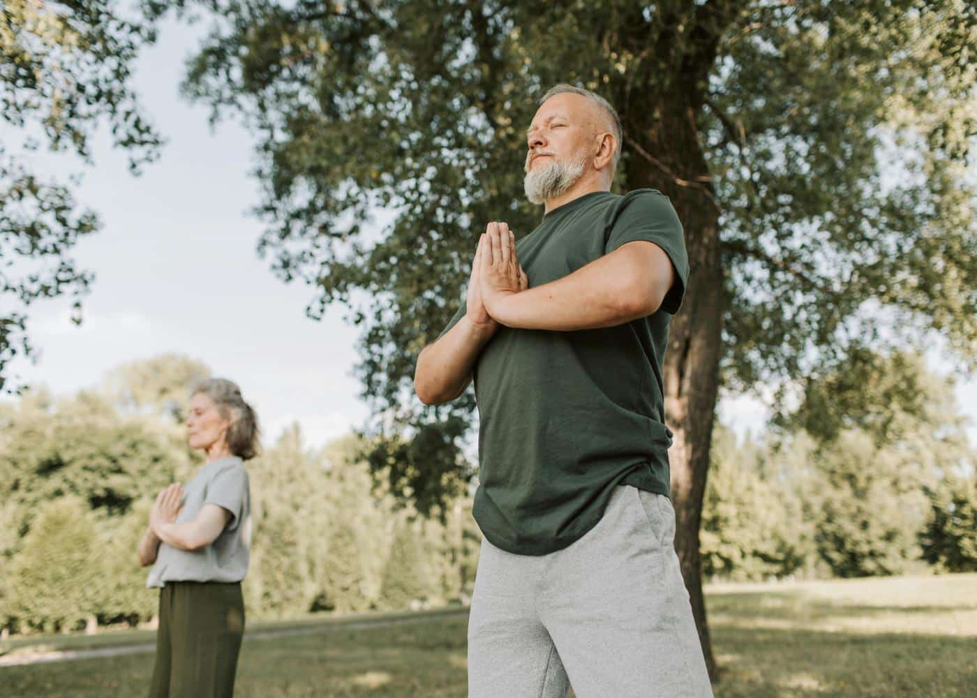 Man outdoors with hands together in front of his chest practicing meditation with a woman doing the same in the background