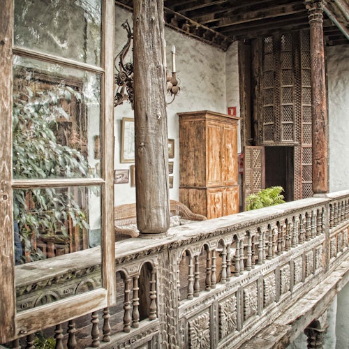 An ornate wooden balcony with detailed railings, rustic furniture, plants, and a hanging chandelier in a vintage setting.