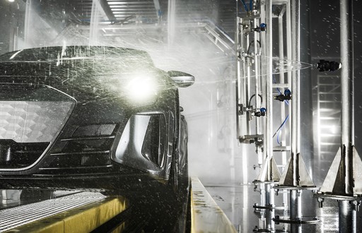 A black car being sprayed with water in an automated car wash, surrounded by metal pipes and nozzles, with headlights glowing through the mist.