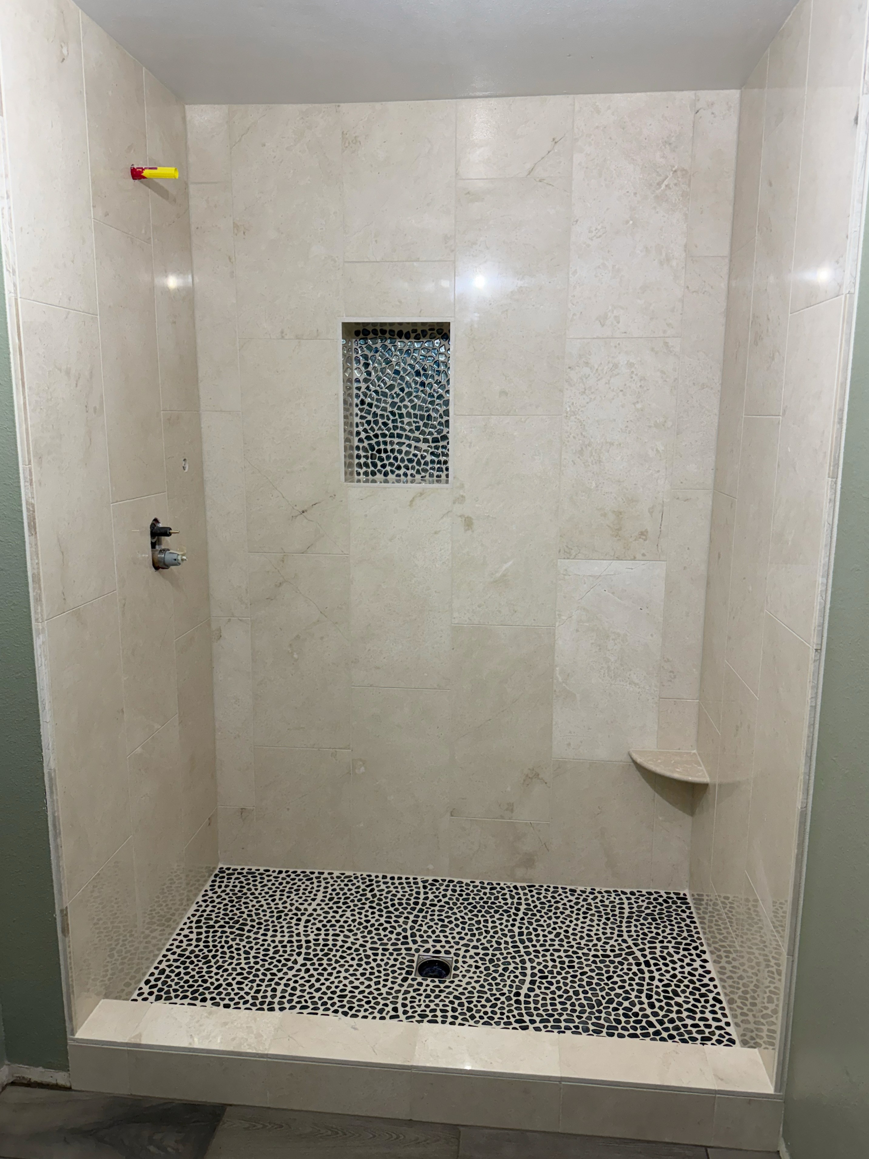 Elegant shower space with light beige natural stone wall tiles, a built-in corner shelf, a custom mosaic tile shower niche, and a black and white pebble mosaic shower floor.