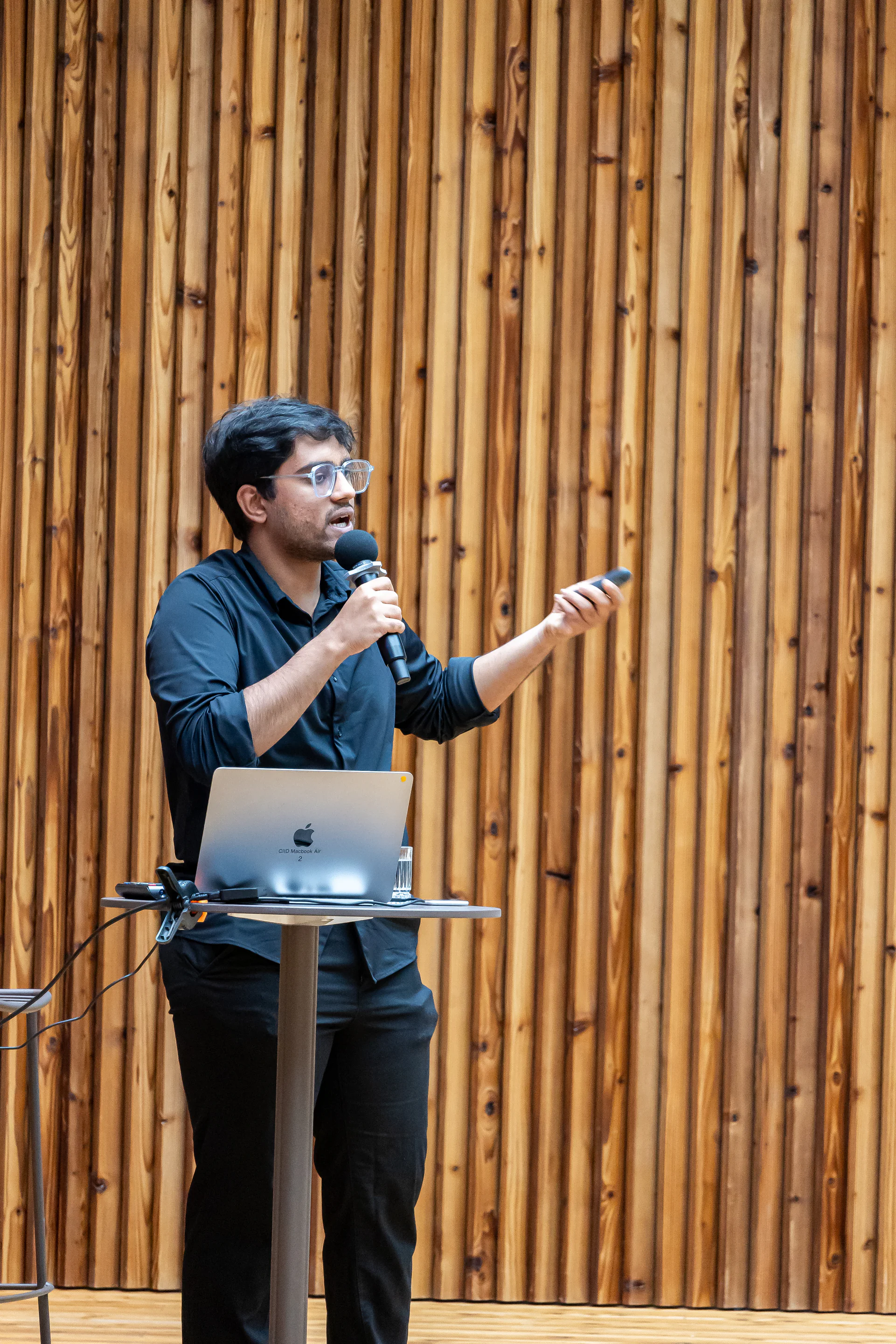 A presenter in black speaks into a microphone beside a laptop against a vertical wood wall.