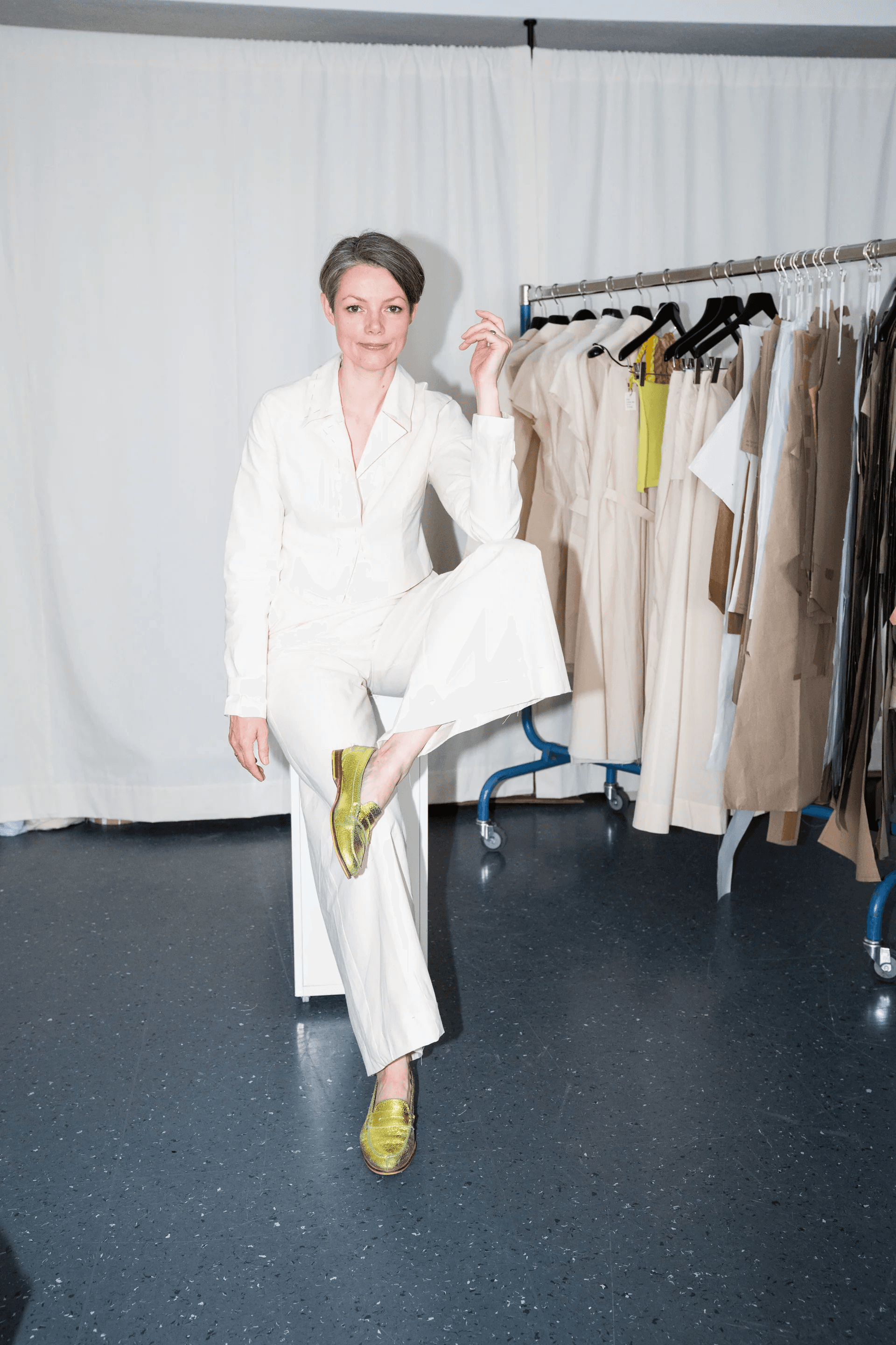 A person in a stylish white pantsuit poses confidently in front of a rack of beige and brown garments, striking a playful pose with one leg bent, and wearing shiny green shoes, set against a minimalist backdrop.