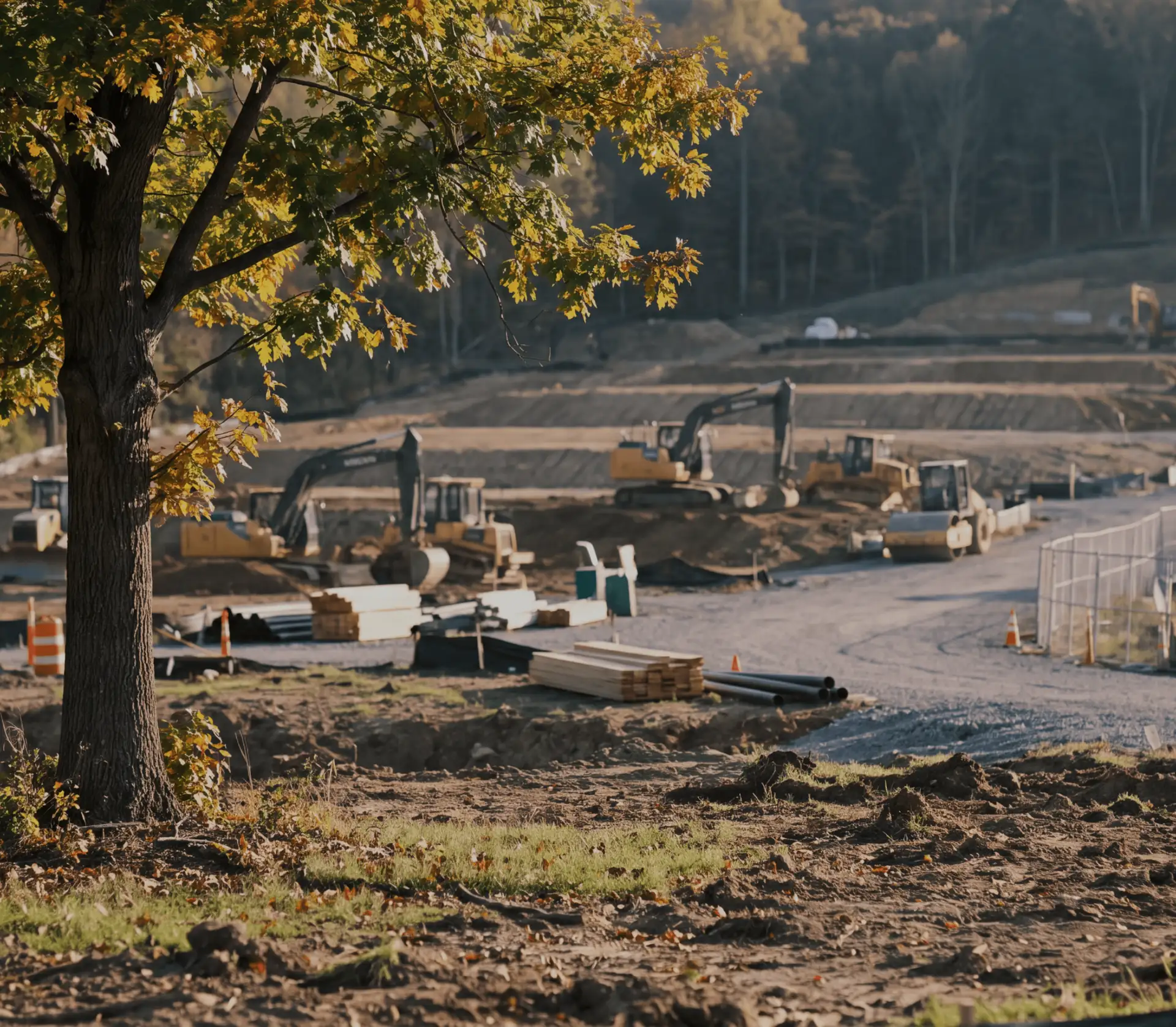 Excavators and a roller grading a cleared hillside for a new subdivision, with stacked lumber and pipe on site and an oak in autumn color in the foreground