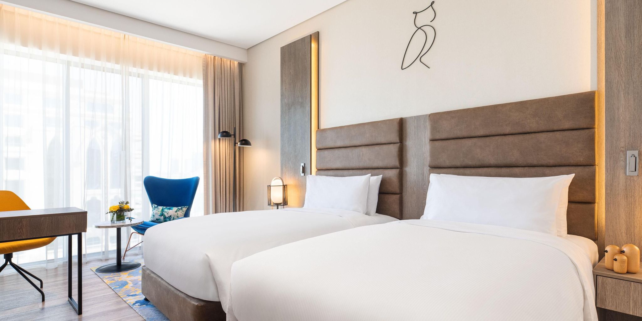 A hotel room in Dubai with two single beds, a desk, chairs, and floor-to-ceiling windows.
