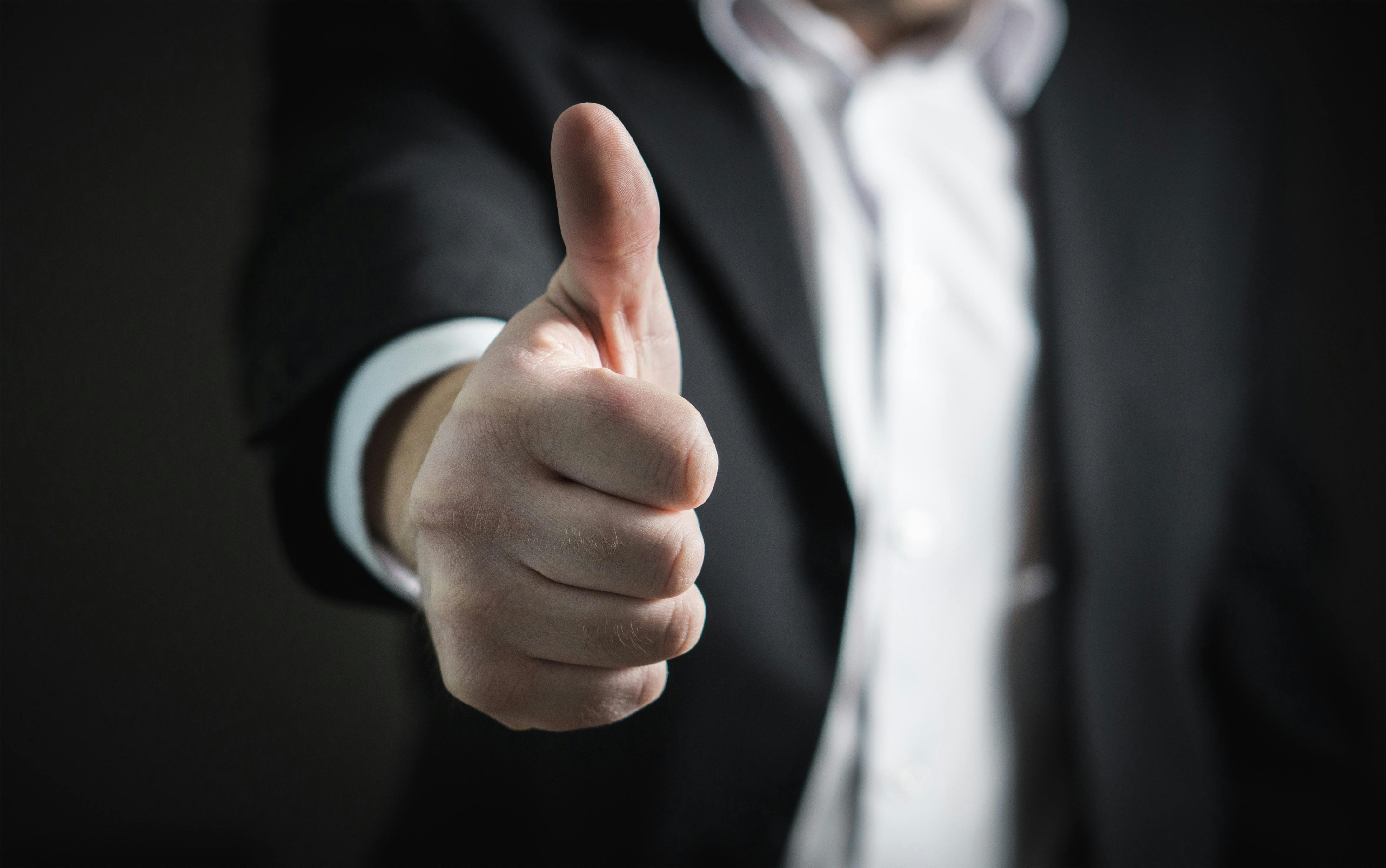 Thumbs up gesture by person in business suit, signifying approval and positivity.