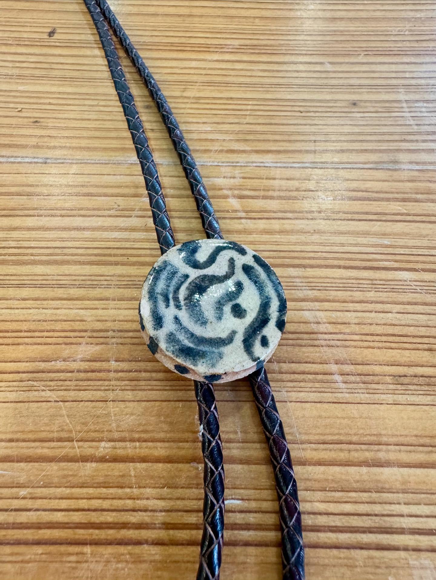 Round ceramic disc with dark blue-green swirls on cream, on black braided cords.