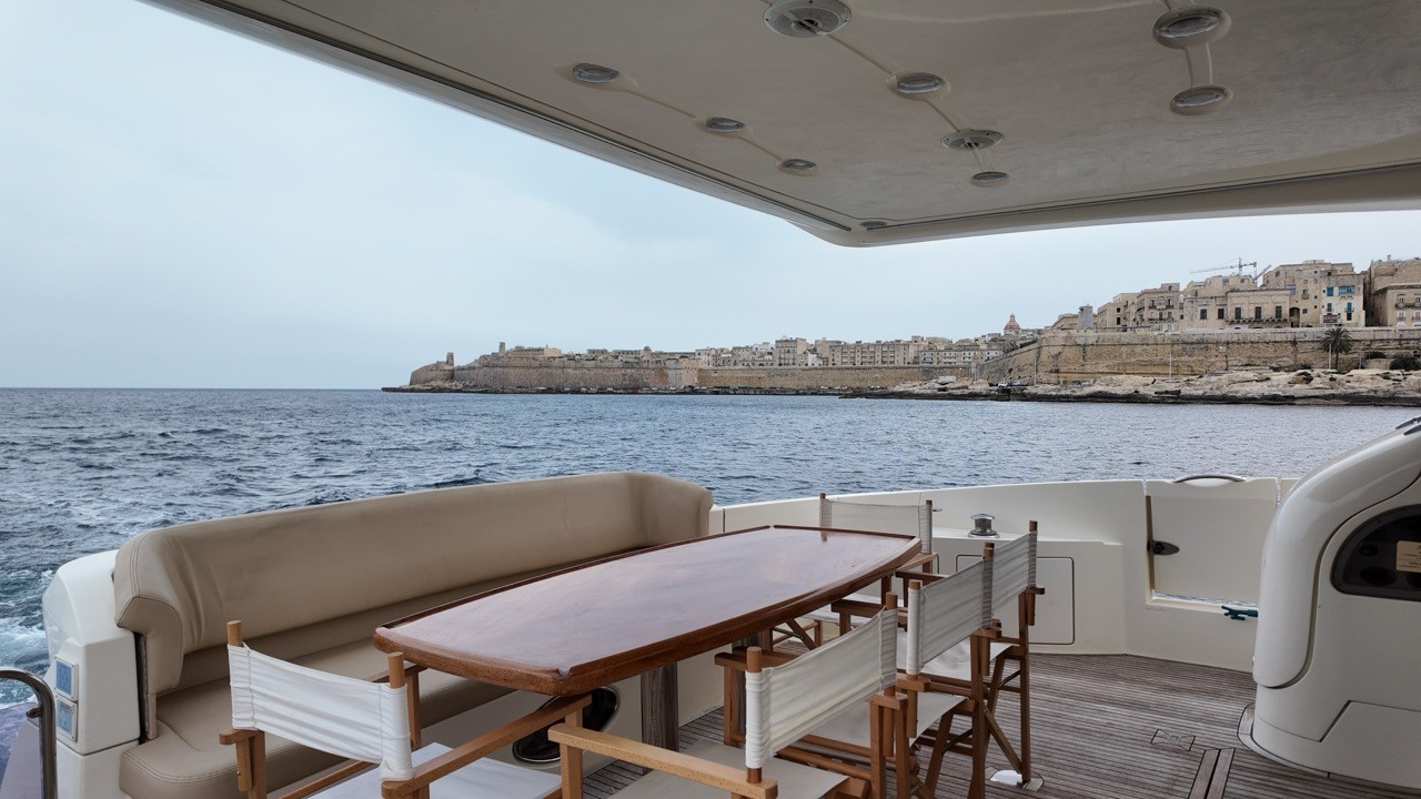 Azimut 68 Evolution aft cockpit dining area with cream leather L-shaped sofa, solid wood dining table, and folding director chairs, sheltered under the flybridge overhang with panoramic views of the Grand Harbour and Valletta skyline across the open sea