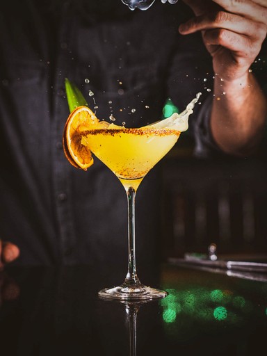 Bartender pouring a yellow cocktail into a martini glass. The drink is garnished with an orange slice and a sprig of mint.