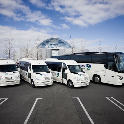 Cuatro furgonetas blancas de transporte y un gran autobús turístico blanco estacionados frente a un edificio moderno con cúpula.