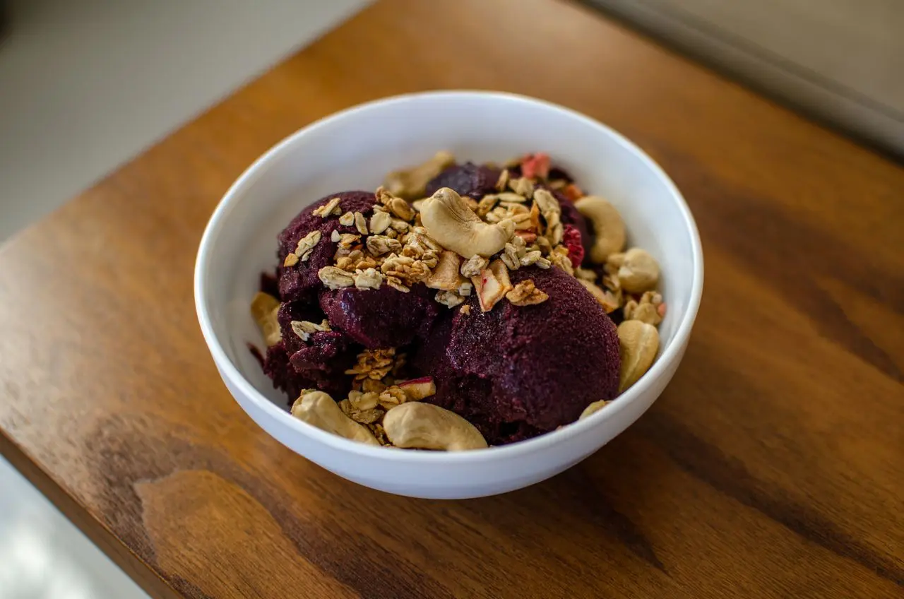 Tigela com sorvete de açaí servido em bolas, coberto com granola, castanhas-de-caju e pedaços de frutas secas, sobre uma mesa de madeira.