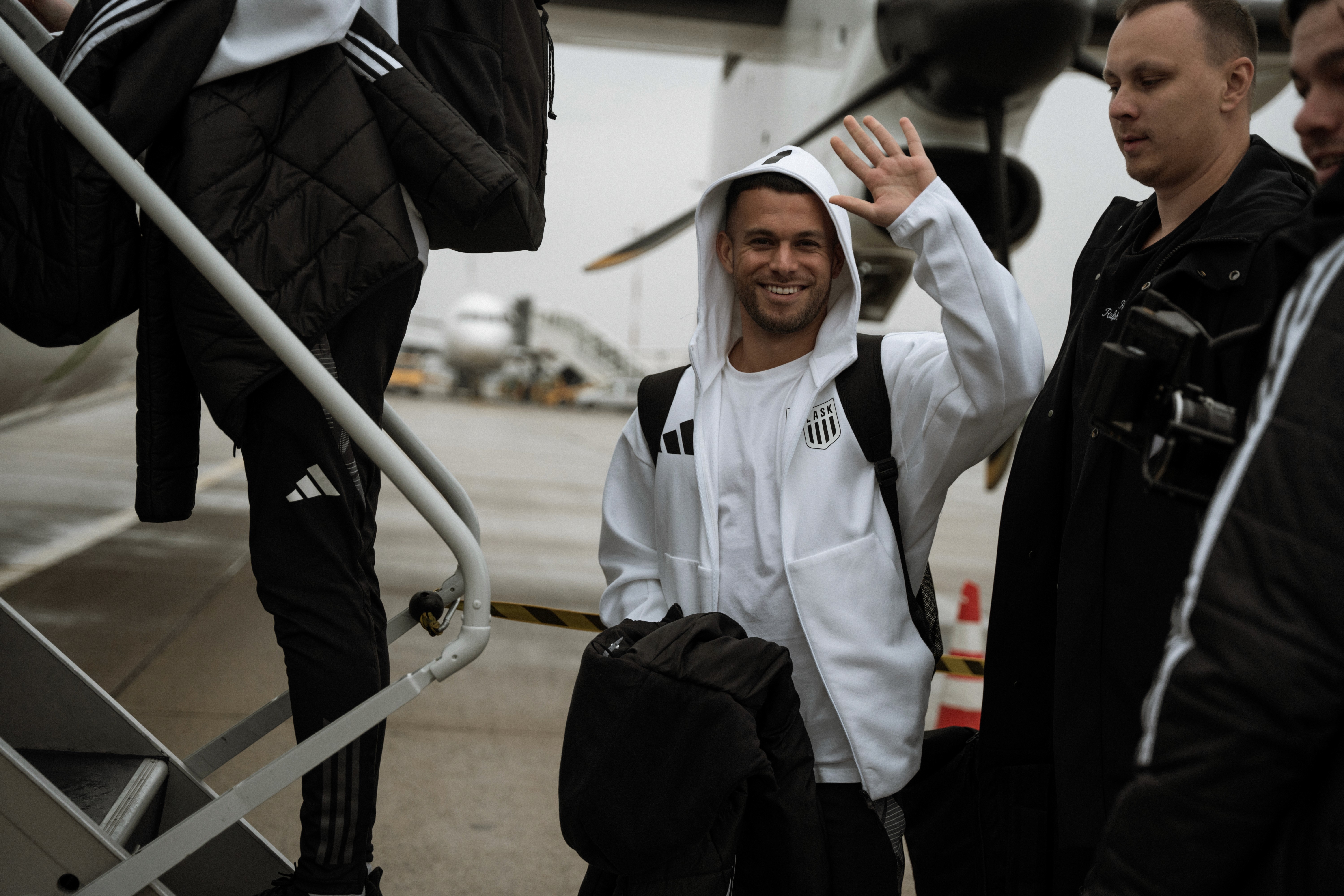 Ein Mann in weißem Kapuzenpullover winkt vor einem Flugzeug. Er trägt einen Rucksack und eine schwarze Tasche.