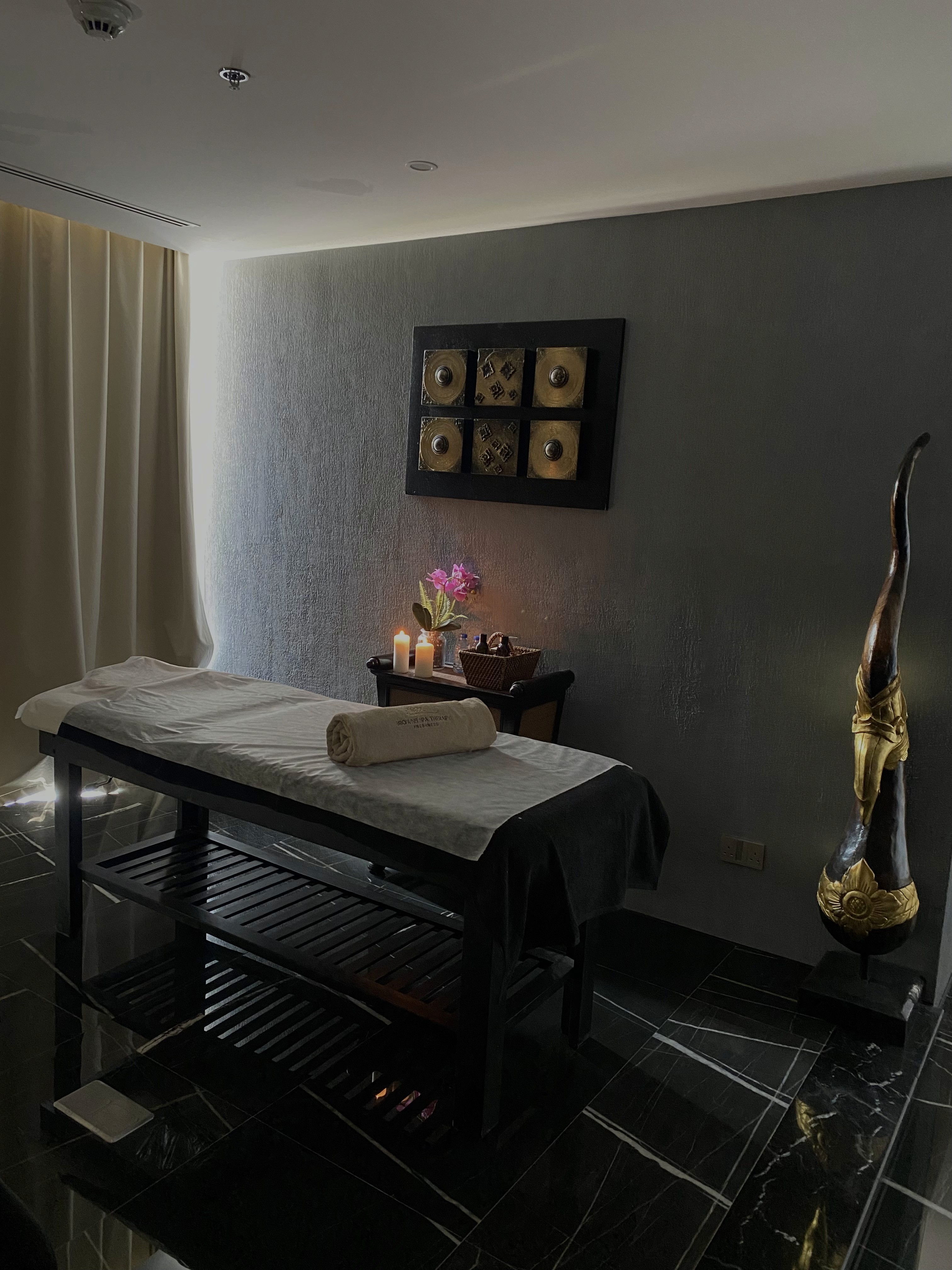 A serene and elegant spa space with wooden reclining chairs and white cushions. Golden sunlight filters through a cascading water feature, creating a peaceful atmosphere. Tropical plants and candles add to the relaxing ambiance.