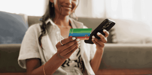 A woman sitting on a couch holds a smartphone in one hand and a credit card in the other, focused on her device.