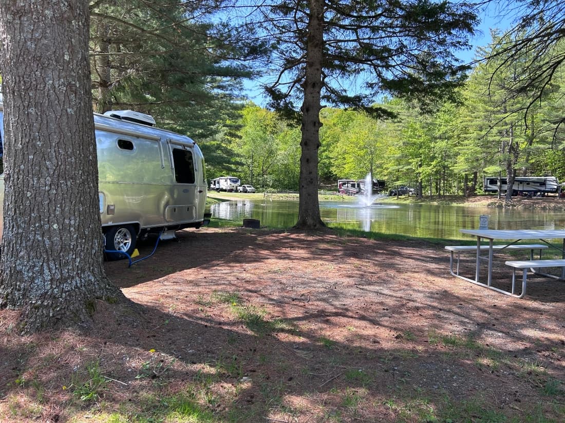 Airstream parked at a shaded waterfront RV site with a picnic table and pond view