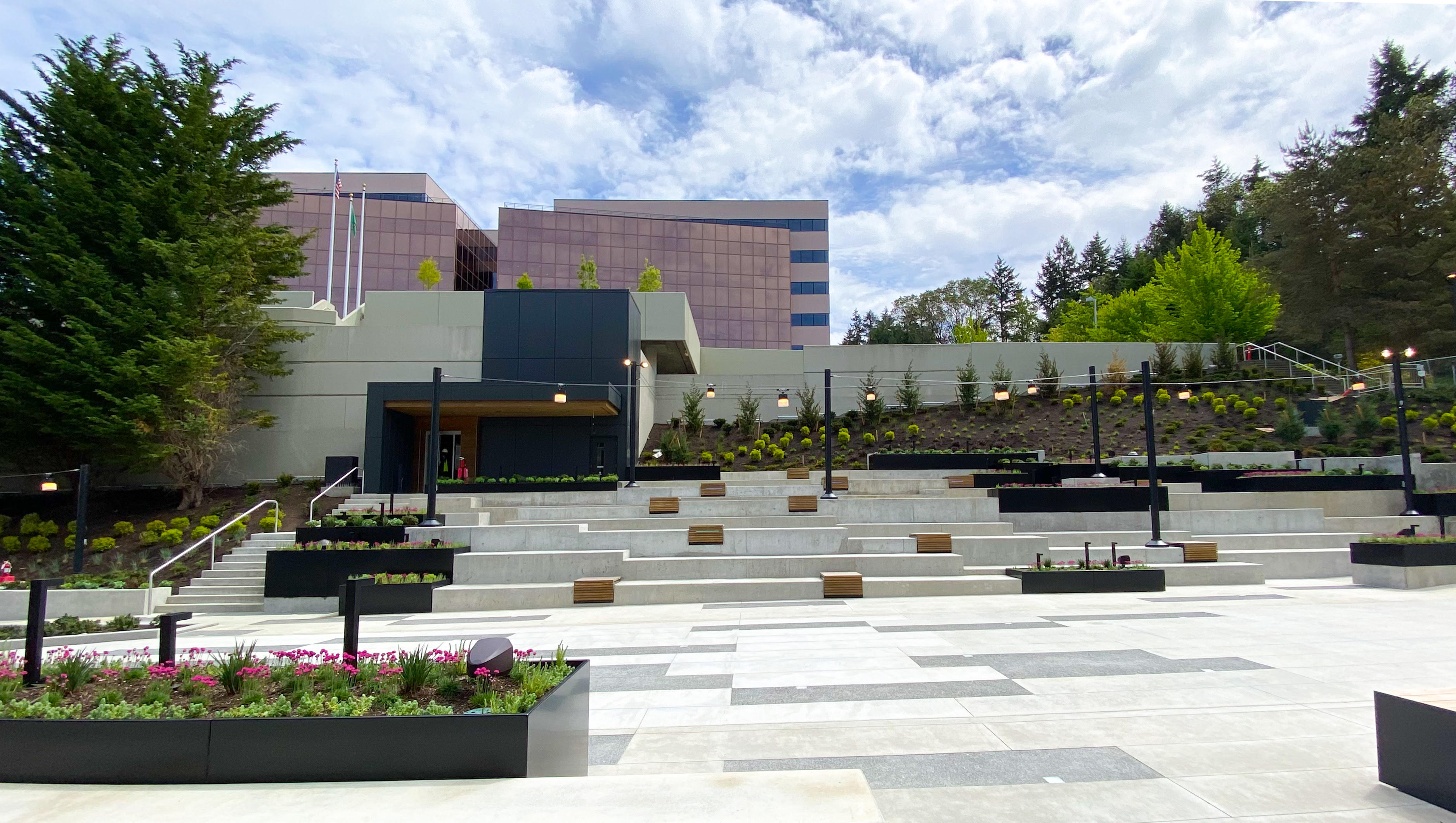 A view across the plaza reveals wooden benches scattered along the concrete amphitheater, while dark metal cladding highlights the pedestrian entrance to the parking garage in the background.