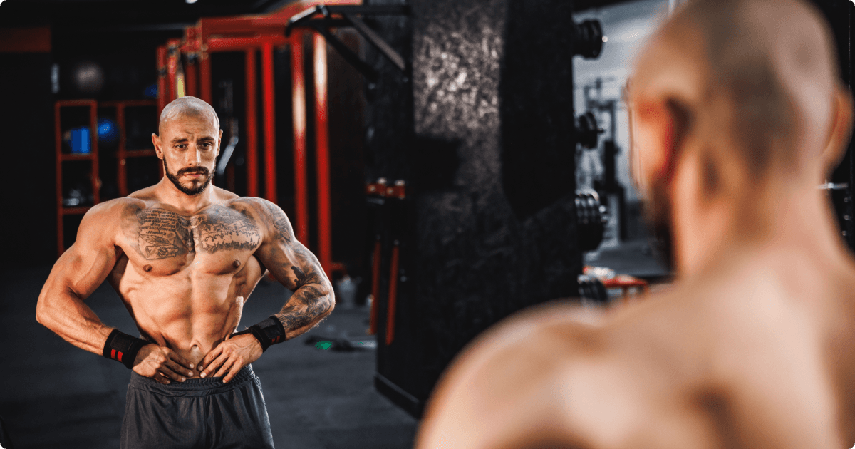 A muscular, tattooed man standing in front of a gym mirror, flexing and assessing his physique