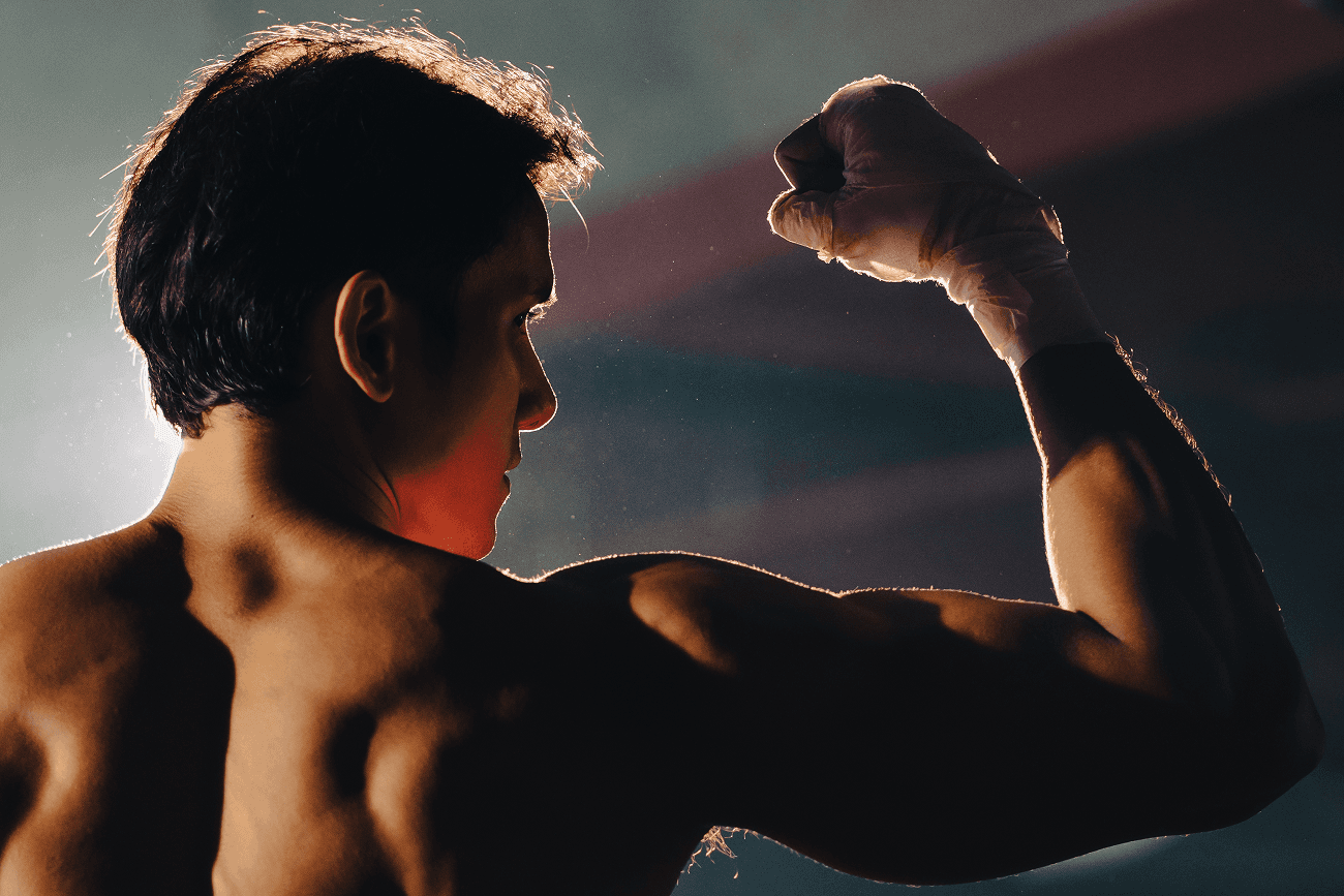 Back view of a muscular man with a wrapped hand flexing his arm under dramatic lighting.