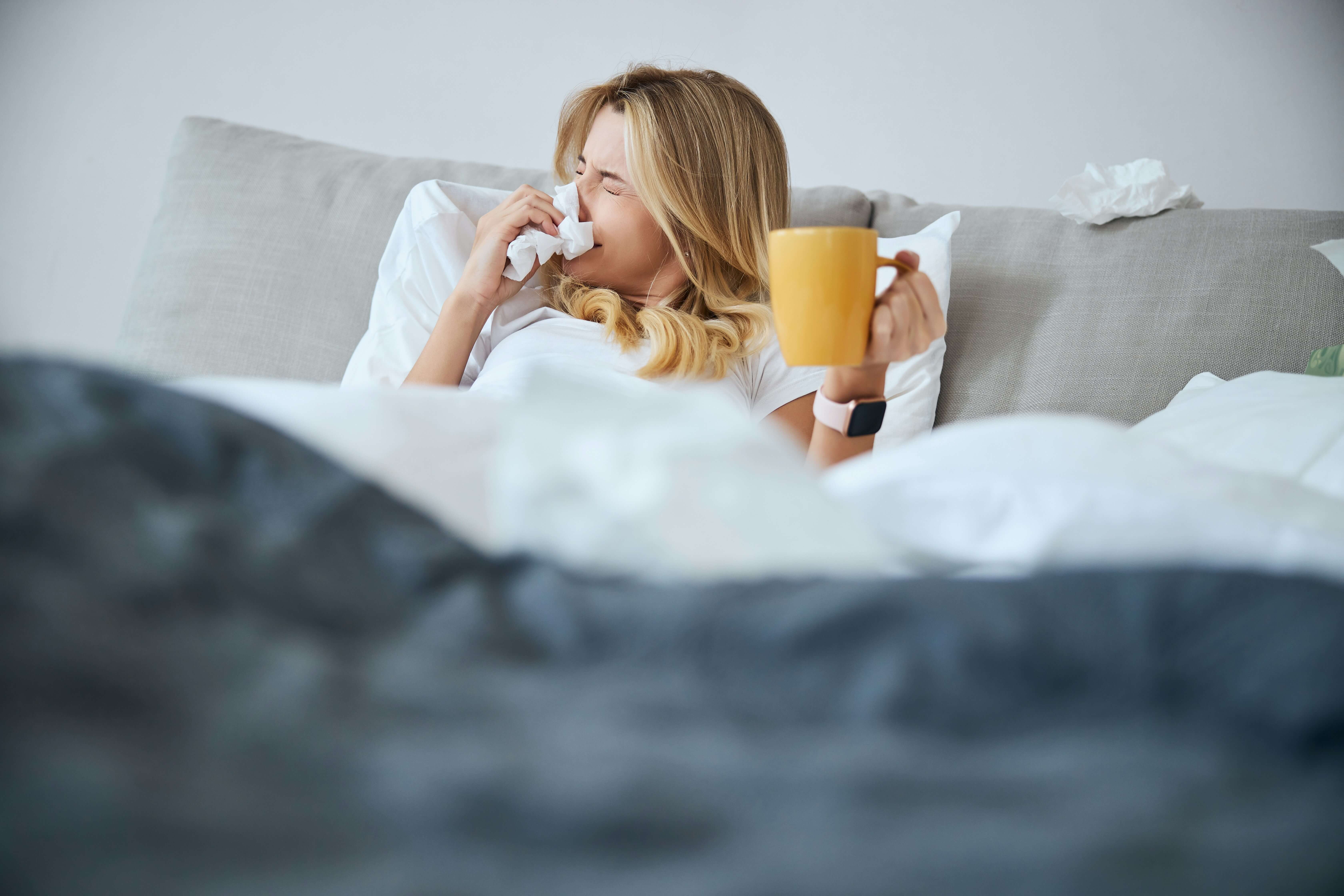 Frau liegt im Bett und schnauft sich die Nase mit einer Tasse Tee in der Hand