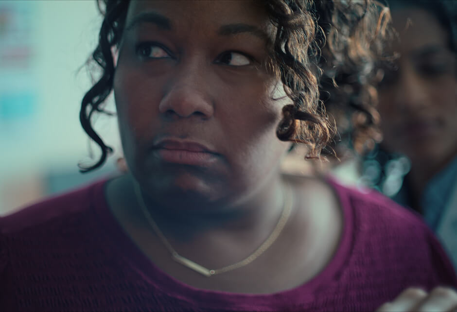 A woman with curly hair and a maroon sweater looks to the side with a serious expression; another person is blurred in the background.