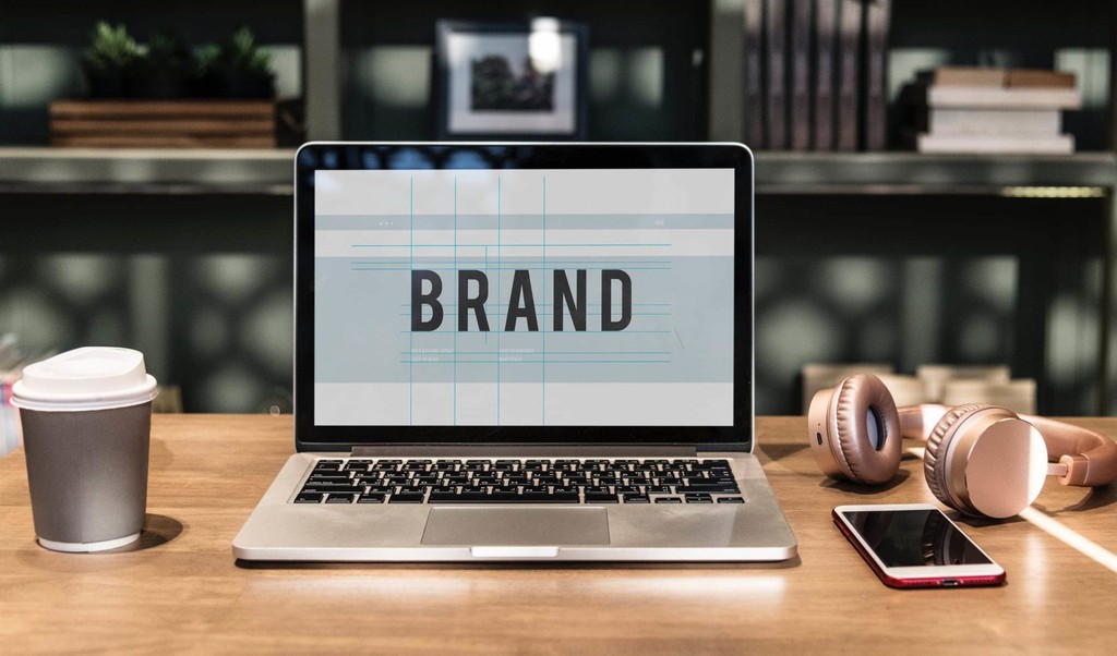 Laptop displaying "brand" with coffee, headphones, and smartphone on a wooden desk.
