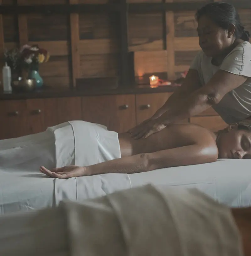 Yäan Spa at Be Tulum: woman relaxing and enjoying a massage, with a masseuse performing techniques, and a lit candle in the background for ambiance.