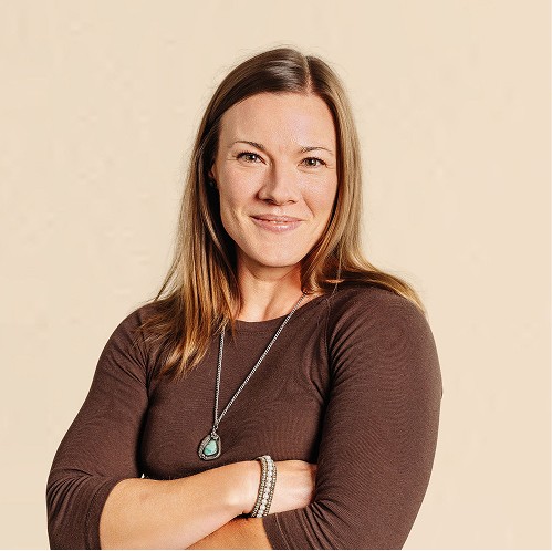 Woman with long hair wearing a brown top and necklace, standing with arms crossed against a neutral background.