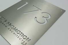 Panneau métallique avec le chiffre « 173 » découpé et les mots « PROMOTION » et « ARCHITECTS » en dessous. Design élégant et moderne ; lettres en relief sur une surface lisse.