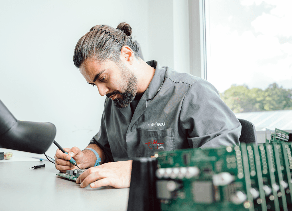 A man in work clothes solders a circuit board, with finished circuit boards visible and blurred in the foreground.