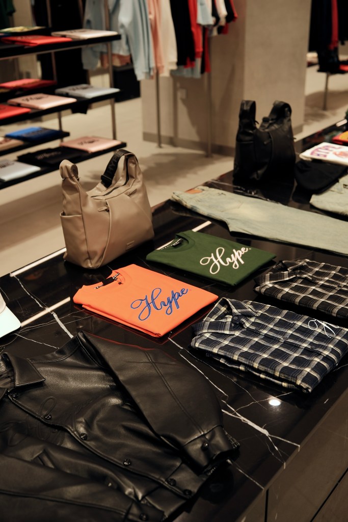 A stylish retail display featuring various clothing items, including a black leather jacket, an orange t-shirt with 'Hype' written on it, a green t-shirt with 'Hype', a plaid shirt, and denim jeans, alongside two handbags.