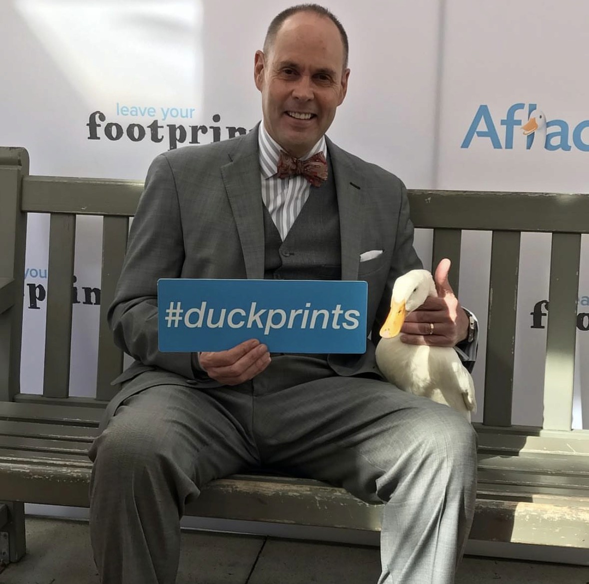 Ernie Johnson in a gray suit sits on a bench, holding the Aflack duck and a blue sign that says #duckprints in white letters. The backdrop features the words leave your footprints and Aflac.