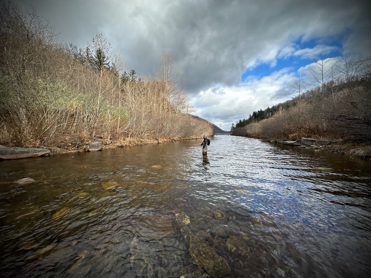Fly angler wading through a wide, rocky river under dramatic skies in late autumn — surrounded by bare trees and distant evergreens in a peaceful mountain valley, capturing the solitude and natural beauty of cold-season fly fishing.