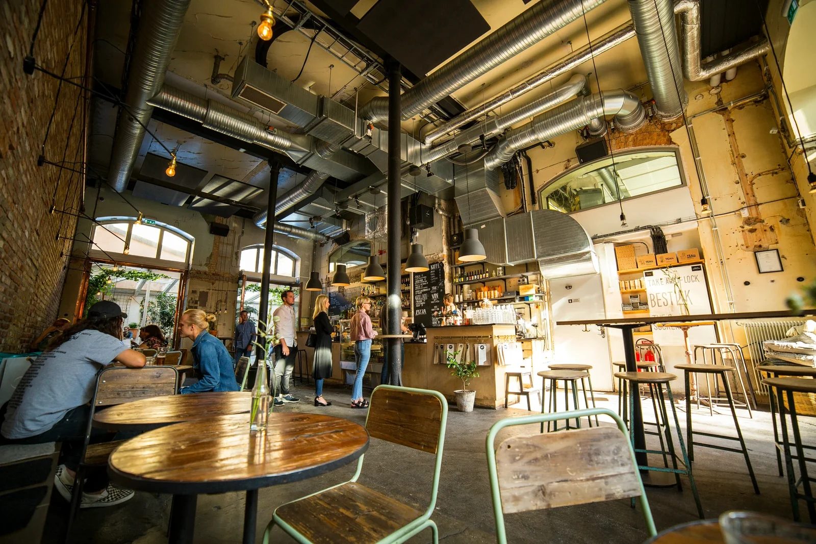 Modern industrial café interior with wooden tables, mint chairs, exposed ductwork, and customers at counter displaying "LOOK BEST" signage.