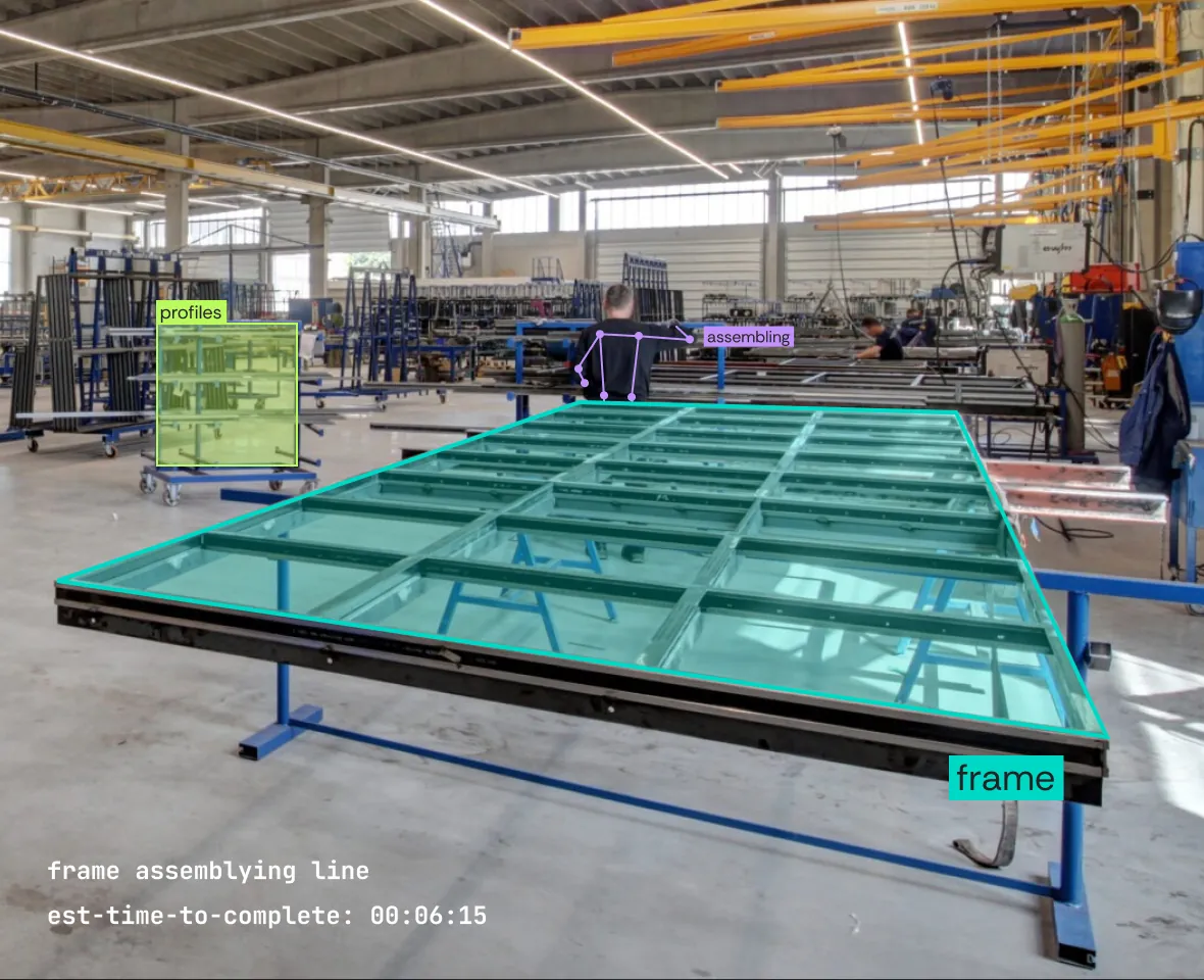 Employee in a bright workshop at JM van Delft assembles a large metal window frame, with overhead cranes and transport trolleys in the background.