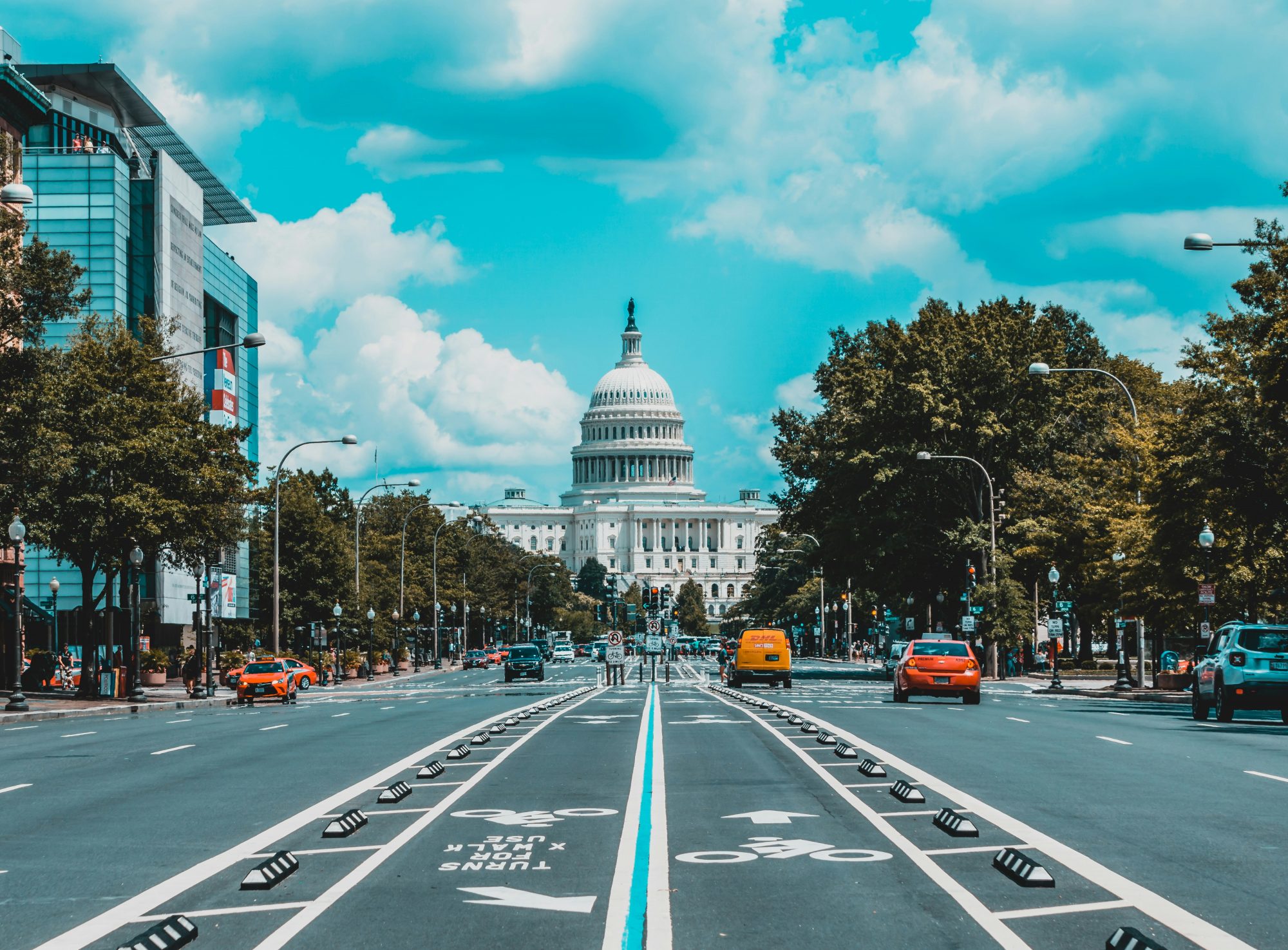 A wide street view leading toward a large dome structure, surrounded by buildings and trees under a blue sky.