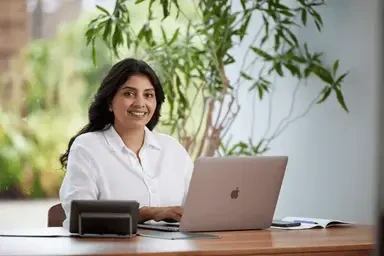 Smiling woman working on a laptop at a wooden desk in a bright office with plants in the background.