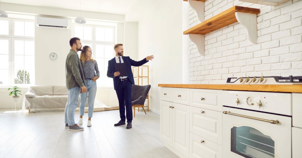 Three people stand in a bright kitchen and living area while one person gestures toward cabinets and shelves.