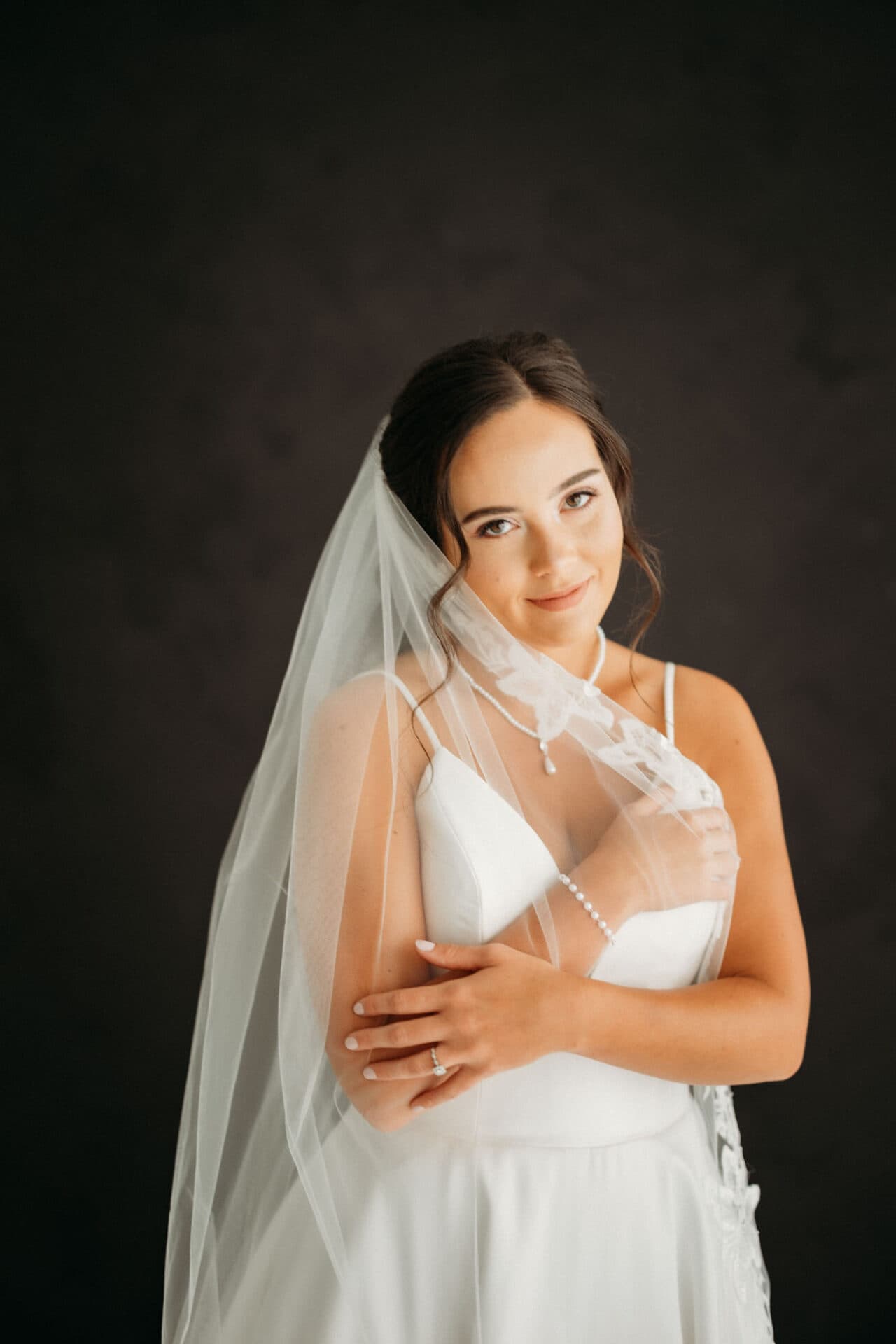 A bride poses with a soft smile and holds her veil against a dark backdrop during a bridal session at Revelator Studio, a natural light studio in Shreveport.